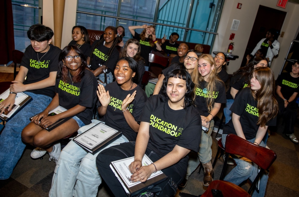 A group of students in Education at Roundabout t-shirts sit in a crowd and smile.